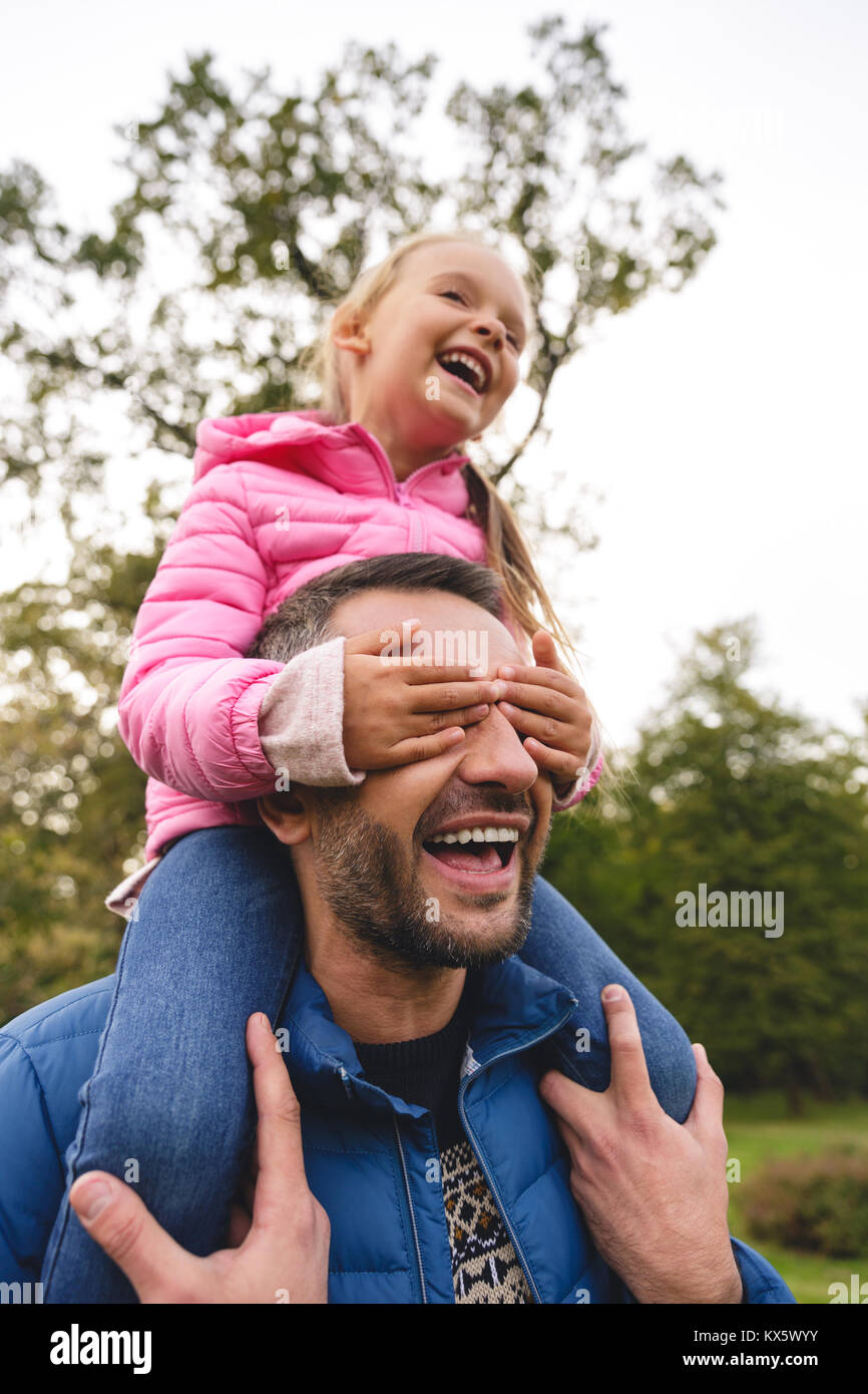 Fröhliches kleines Mädchen reiten auf Vater zurück in den Park, das Mädchen die Augen ihres Vaters deckt Stockfoto