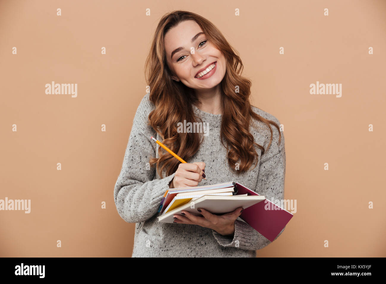 Fröhliche brunette Mädchen in Form gestrickter Pullover schreiben Notizen auf ihrem Skizzenblock während Kamera suchen, auf beige Hintergrund isoliert Stockfoto