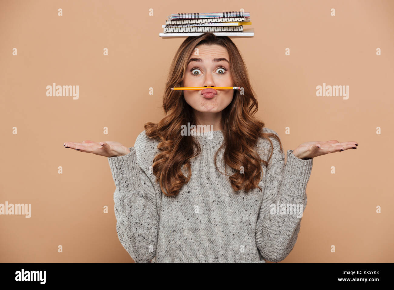 Nahaufnahme von lustig Junge brünette Frau mit Bücher auf dem Kopf halten Bleistift als Schnurrbart und zeigt leere Palmen, auf beige backgroun isoliert Stockfoto