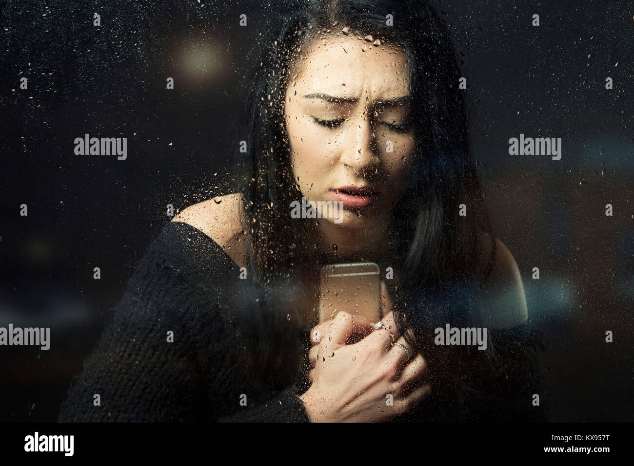 Frau unglücklich, traurig, deprimiert umklammerte Handy geschlossenen Augen weinen suchen verzweifelt sitzen an regnerischen Fenster filmische Nahaufnahme portrait Stockfoto