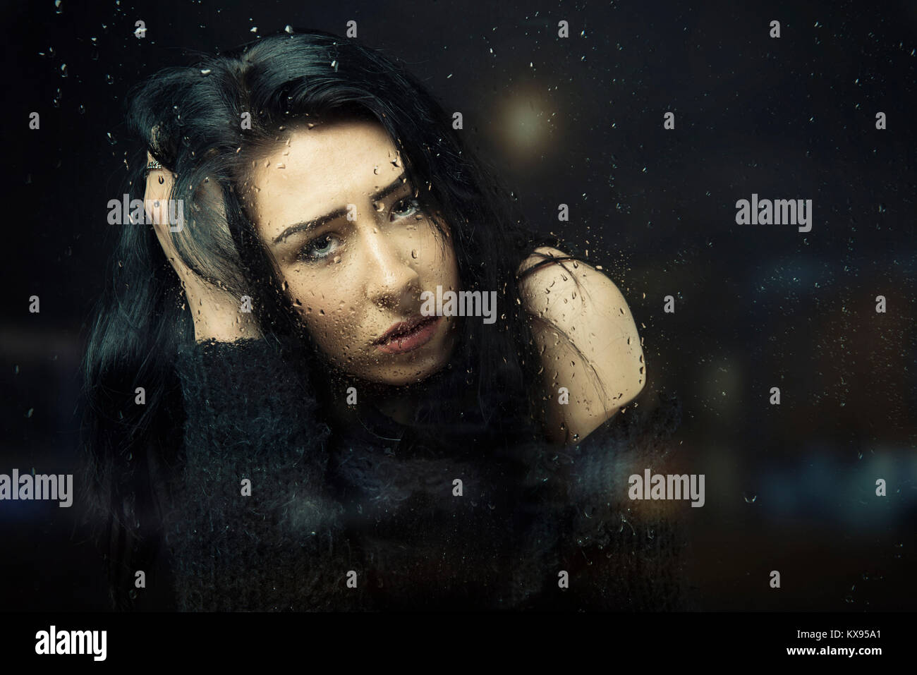 Junge Frau traurig, unglücklich, geradeaus Hand am Haar an regnerischen Fenster filmische Nahaufnahme Portrait sitzen Stockfoto