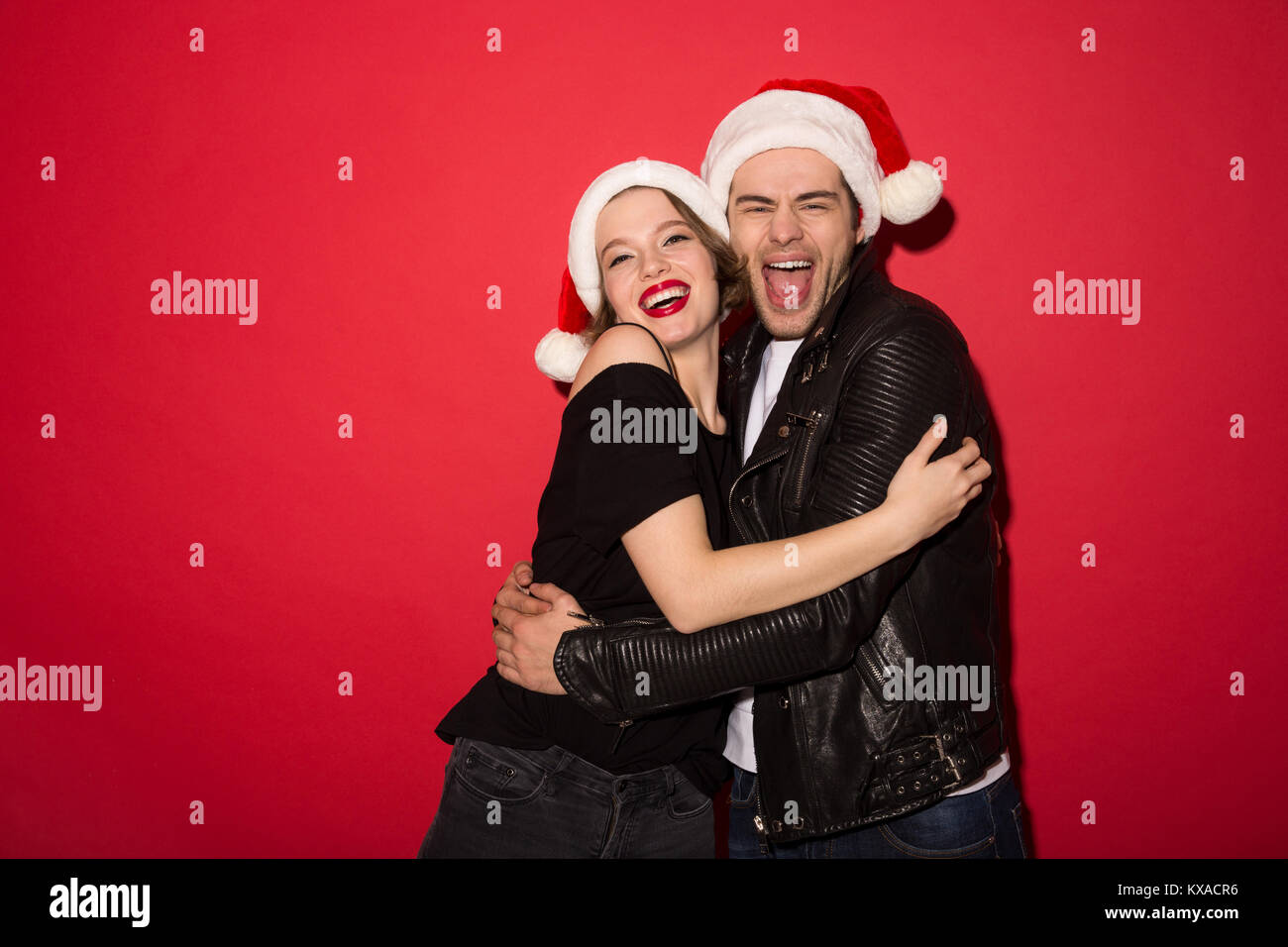 Fröhlichen punk Paar in Weihnachten Hüte umarmen und mit Blick auf die Kamera über den roten Hintergrund Stockfoto