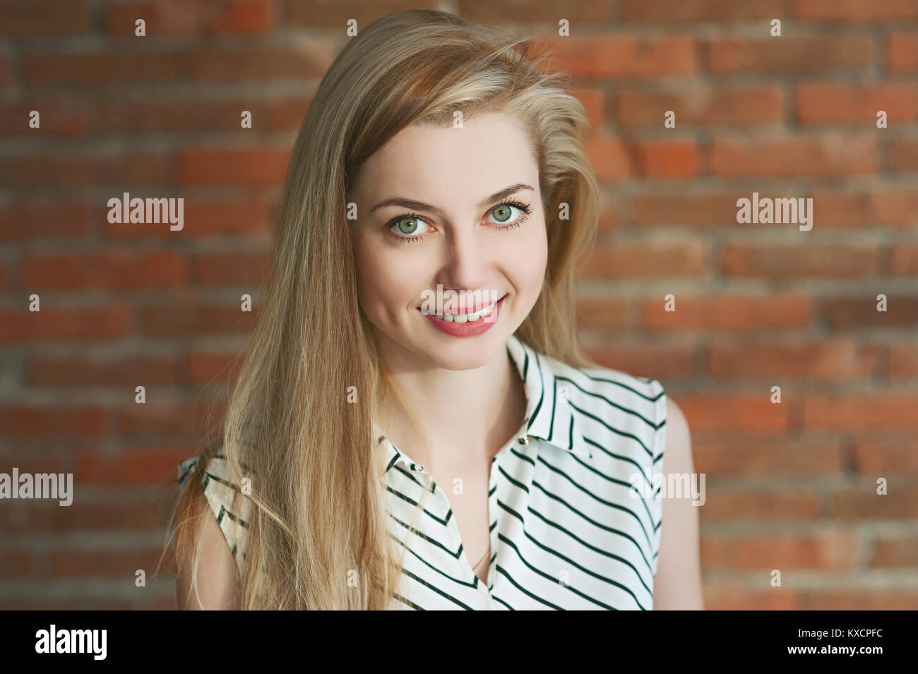 Porträt einer Blondine in ein Licht. Gegen einen Red brick wall Hintergrund, lächelnd und für die Kamera posieren. ihr langes Haar passt in eine Richtung. Stockfoto