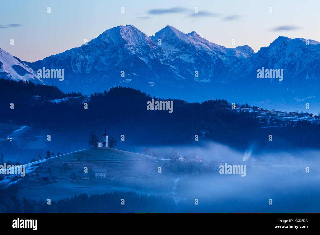 Sicht in der Dämmerung im Winter von Rantovše Hill über zu Sveti Tomaž nad Praprotnim (Kirche des Hl. Thomas) und die Steiner Alpen, Slowenien. Stockfoto