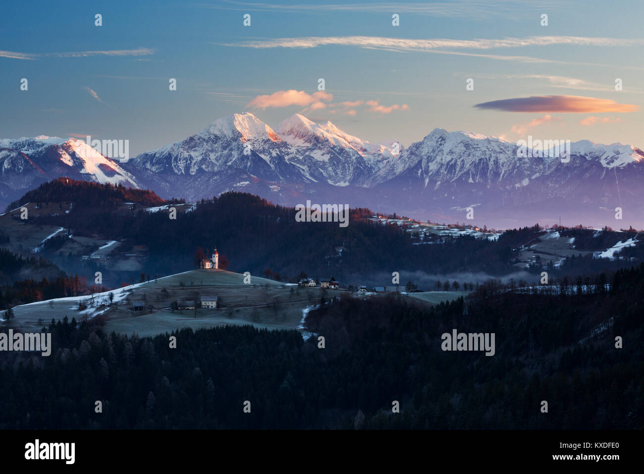 Ansicht bei Sonnenaufgang im Winter von Rantovše Hill über zu Sveti Tomaž nad Praprotnim (Kirche des Hl. Thomas) und die Steiner Alpen, Slowenien. Stockfoto