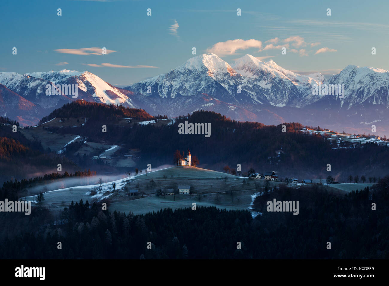 Ansicht bei Sonnenaufgang im Winter von Rantovše Hill über zu Sveti Tomaž nad Praprotnim (Kirche des Hl. Thomas) und die Steiner Alpen, Slowenien. Stockfoto