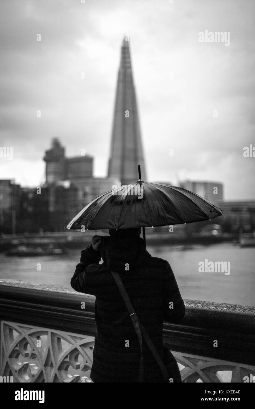 Eine Person, die Schilde sich vom Regen, während sie in Richtung der Shard von der Tower Bridge über die Themse in London Stockfoto