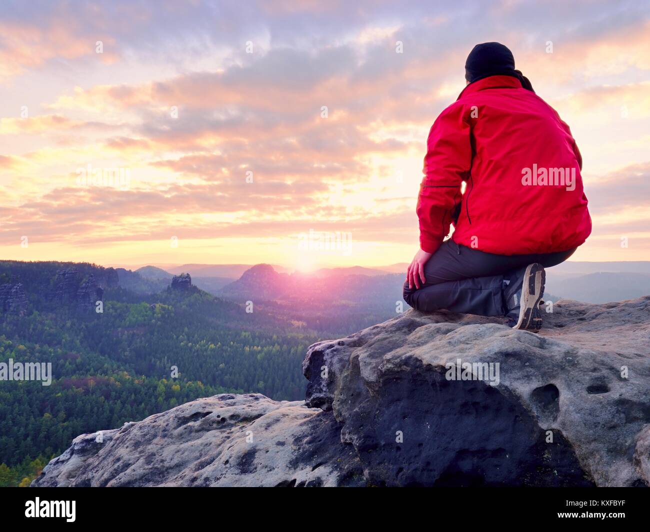 Sitzung Wanderer auf Rock. Mann in Rot Schwarz warme Kleidung sitzt auf Felsen und weiten Blick genießen. Blick in Misty hügeligen Tal unten Stockfoto