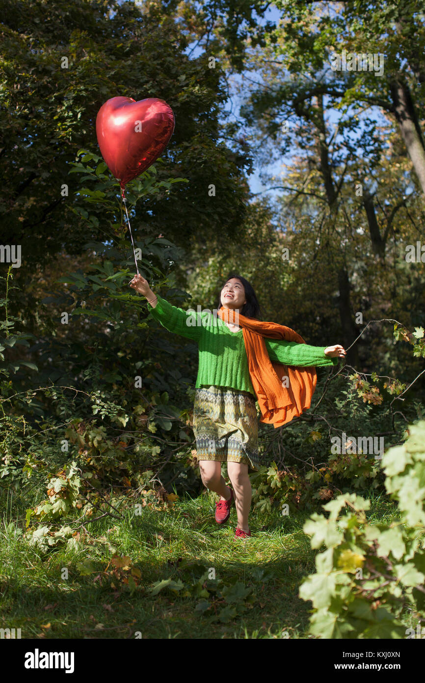 Glückliche junge Frau hält einen roten Herzballon, während sie im Park gegen Bäume läuft Stockfoto