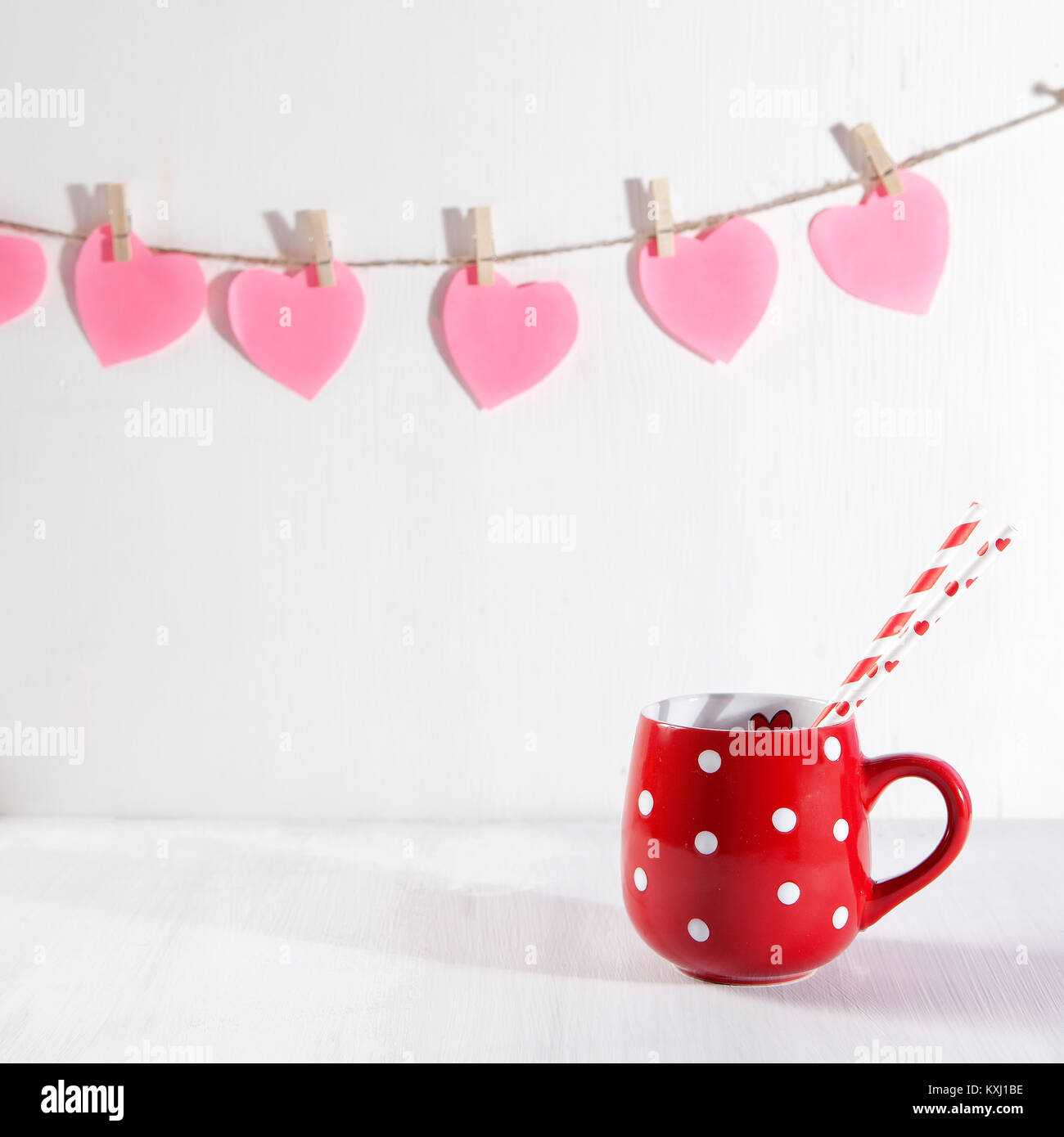Red Cup, der mit einem weißen Punkt auf einem weißen Holztisch. Papier Herzen hängen an der Wand Stockfoto