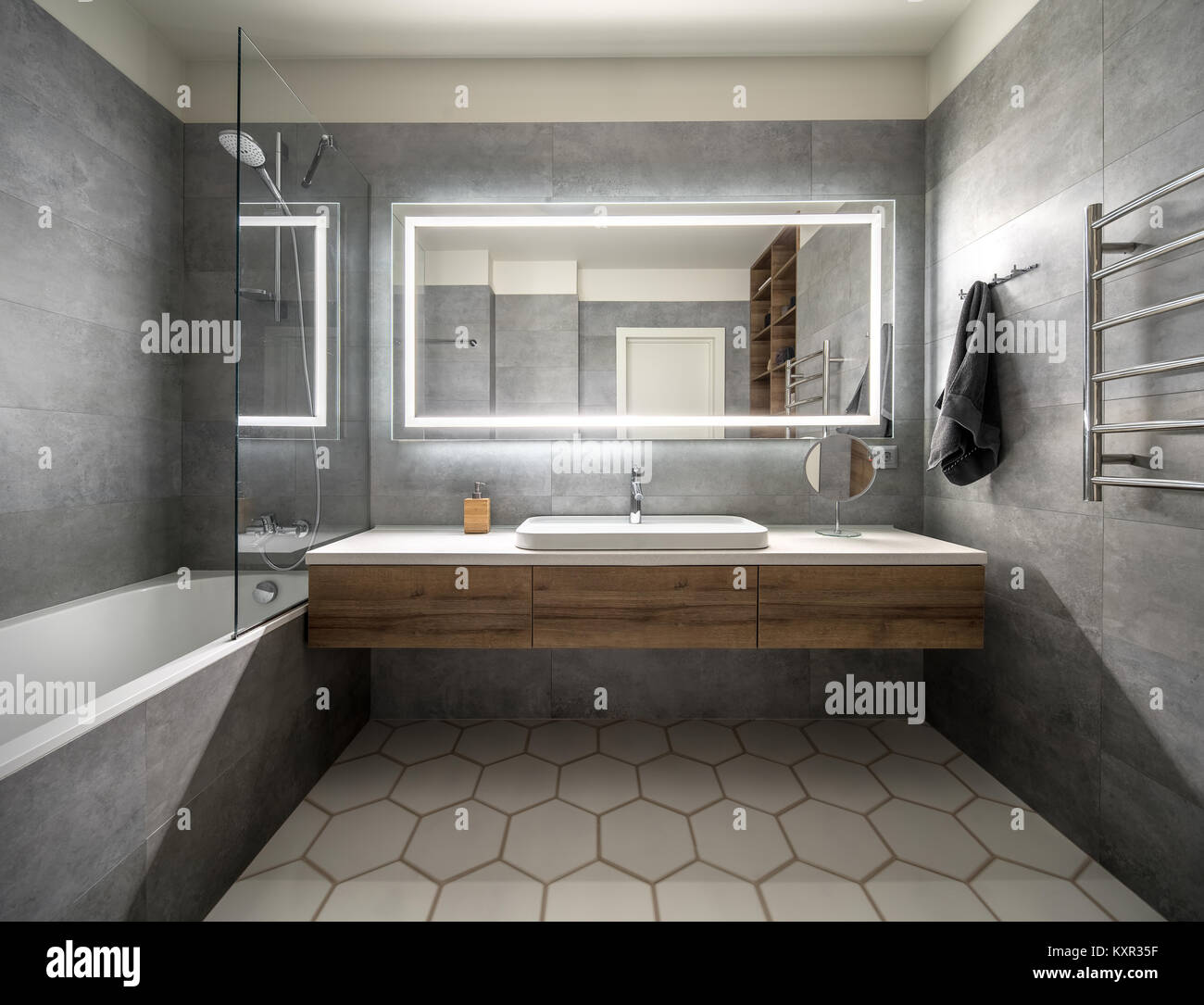 Badezimmer in einem modernen Stil mit grauen und weißen Fliesen. Es gibt einen großen Spiegel mit leuchtenden Lampen, Tischplatte mit holzschubkästen und Waschbecken, Badewanne mit sh Stockfoto