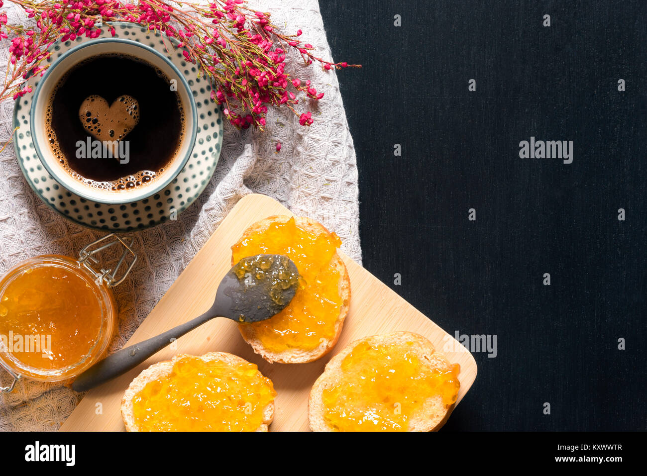 Frühstück mit einer Tasse Kaffee mit einem Schaum Herz, Brotscheiben mit Pfirsich Marmelade auf einem Schneidebrett, umgeben von Blumen verschmiert, auf einem Schwarzen Tisch. Stockfoto