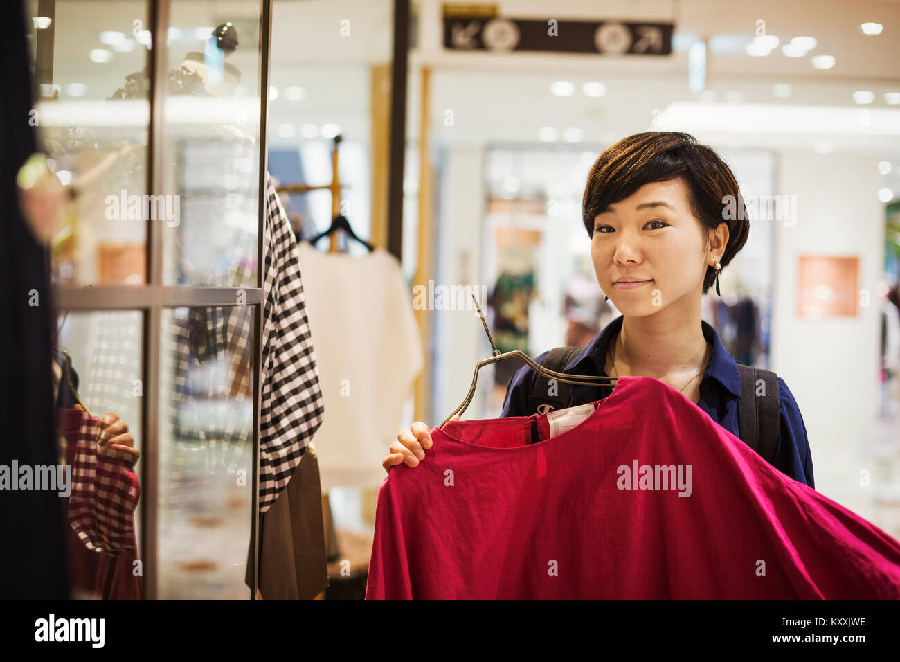 Frau mit schwarzen Haaren trägt blaue Hemd steht drinnen, an Kleidung in einem Geschäft suchen, lächeln in die Kamera. Stockfoto