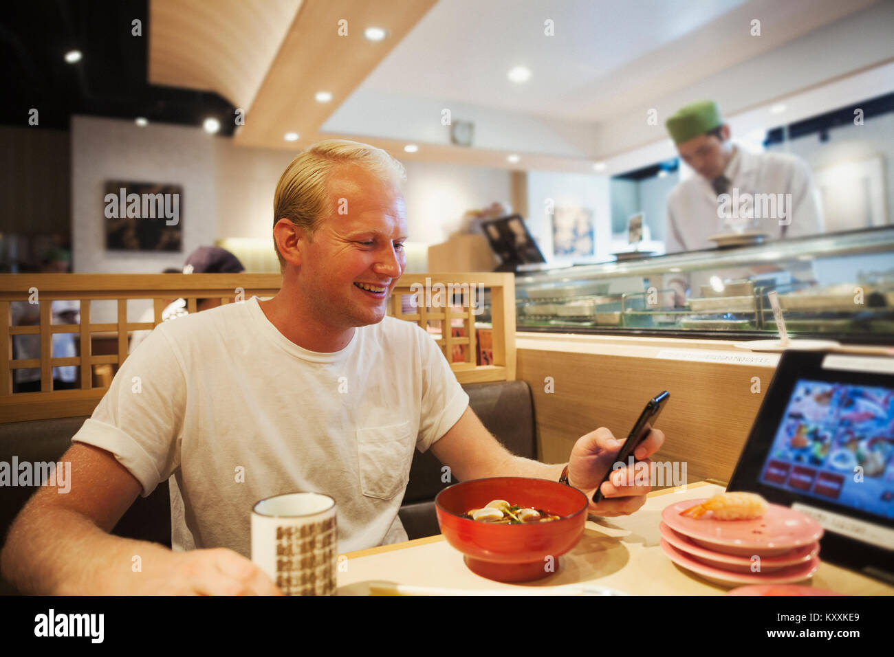Junger Mann an einem Tisch in einem asiatischen Fast Food Restaurant zu sitzen, bei Smartphone suchen, lächelnd. Stockfoto
