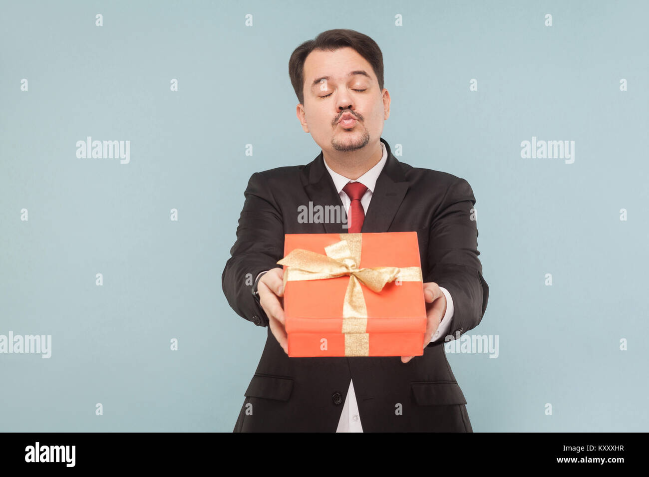 Glücklicher Mann im schwarzen Anzug halten und küssen und roter Geschenkbox. Studio shot, auf hellblauem Hintergrund isoliert Stockfoto