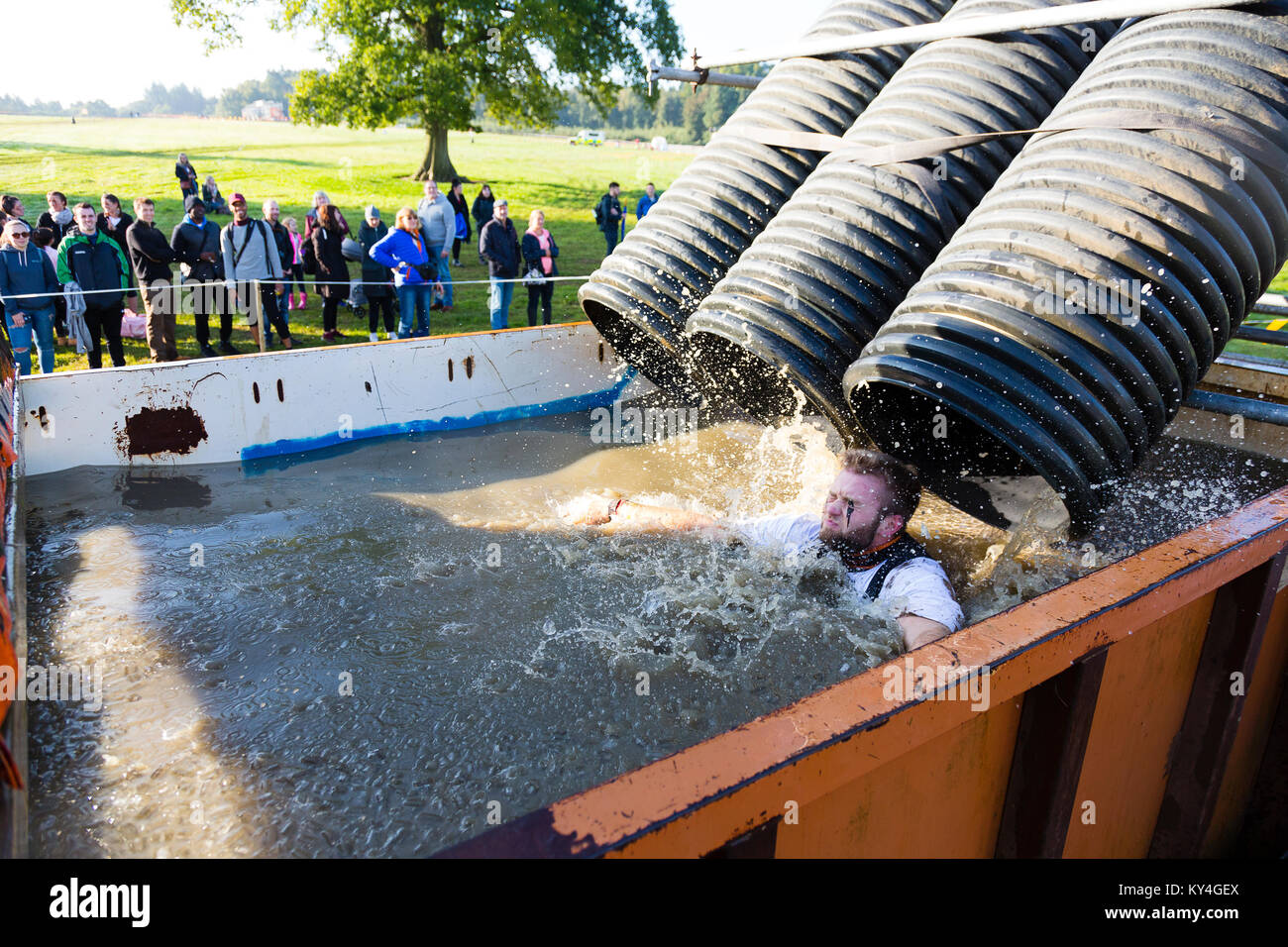 Sussex, UK. Eine Gruppe von Zuschauern beobachten, wie ein Mann nach unten rutschen eine schwarze Plastik Schlauch in einen großen Pool von eiskalt Wasser während einer haltbaren Mudder Ereignis Stockfoto