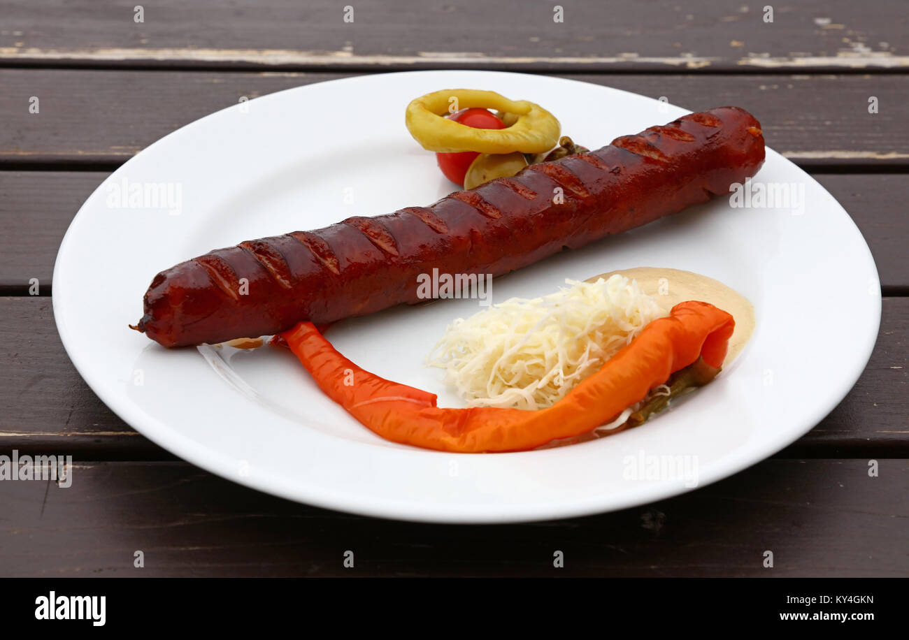 Close up Teil der geräucherte Bratwurst mit Beilage von eingelegtem Pfeffer, Meerrettich und Senf auf weißen Platte über braune Holztisch, hohe Angl Stockfoto