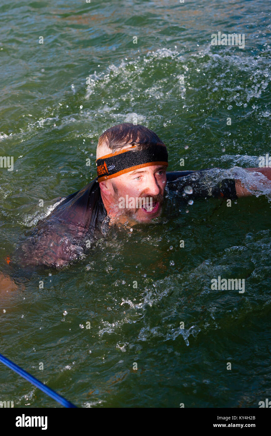 Sussex, UK. Ein männlicher Mitbewerber schwimmt an der Seite, nachdem Sie in das Wasser auf harten Hindernis der Hangin' während einer haltbaren Mudder Ereignis. Stockfoto