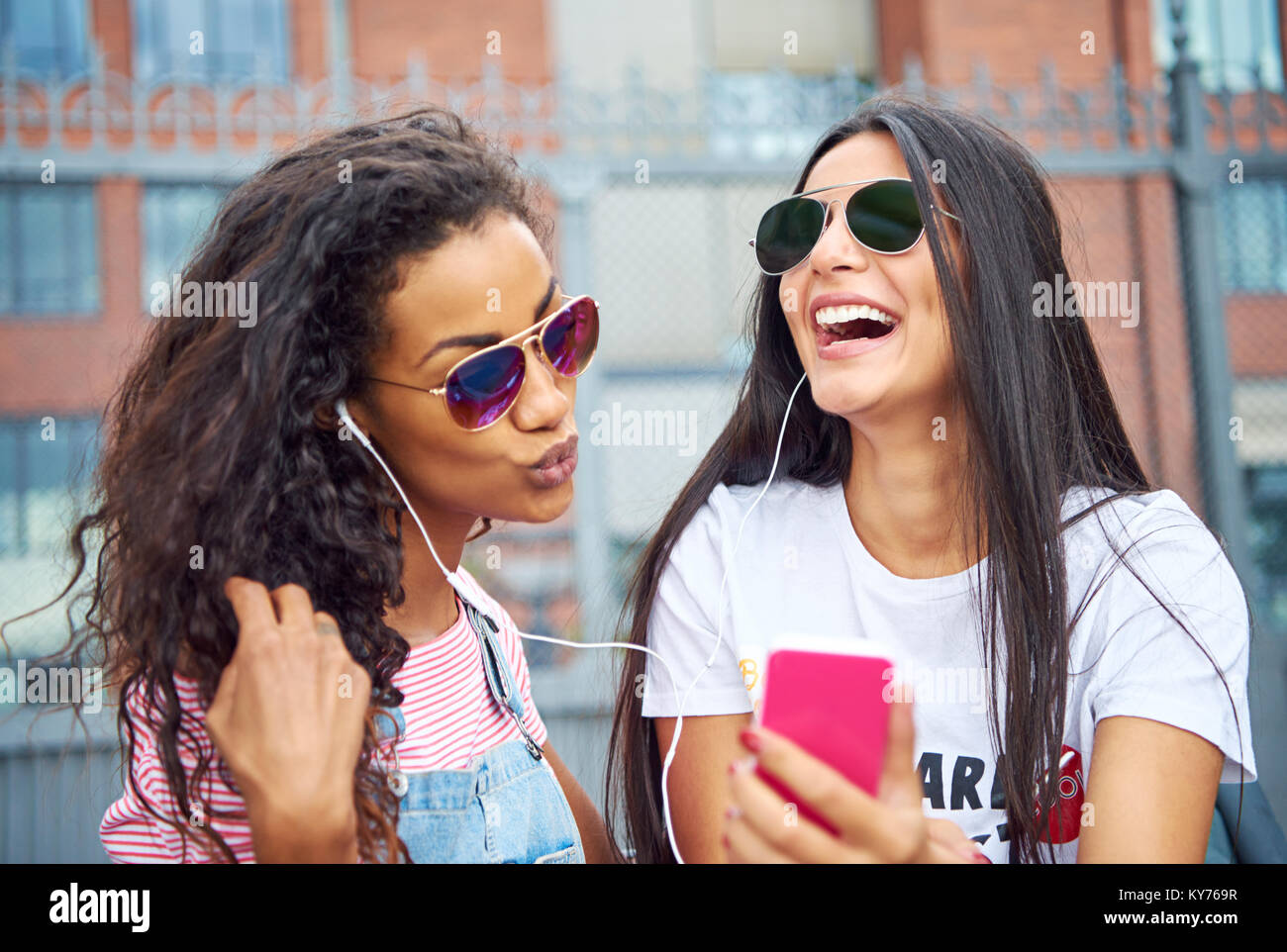 Zwei Lachende junge Freundinnen sitzen auf einem City Bank zu Musik über Kopfhörer und Selbstporträts zusammen Hören mit einem Smartphone Stockfoto