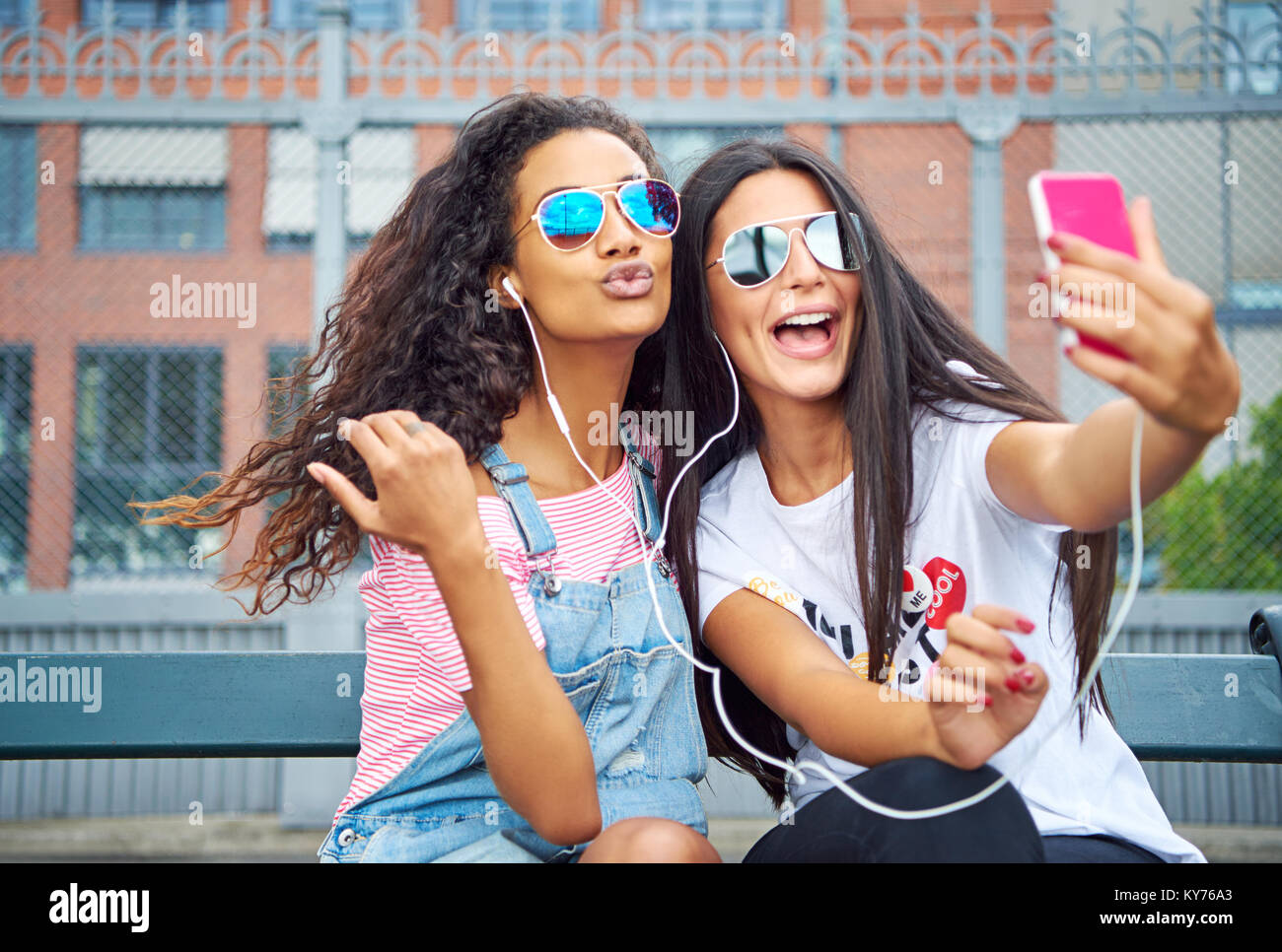 Zwei junge Freundinnen lächelnd und sitzt auf einem City Bank zu Musik über Kopfhörer und Selbstporträts zusammen Hören mit einem Smartphone Stockfoto