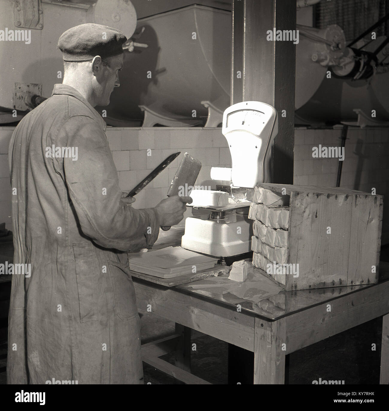 1950, historische, männlichen Arbeitnehmer in Mantel und Kappe mit einem dünnen Metall Spatel und Paddel aus Holz in seinen Händen wiegen ein Pat der Butter auf eine Waage, bevor es im Papier, Irland gewickelt ist. Stockfoto