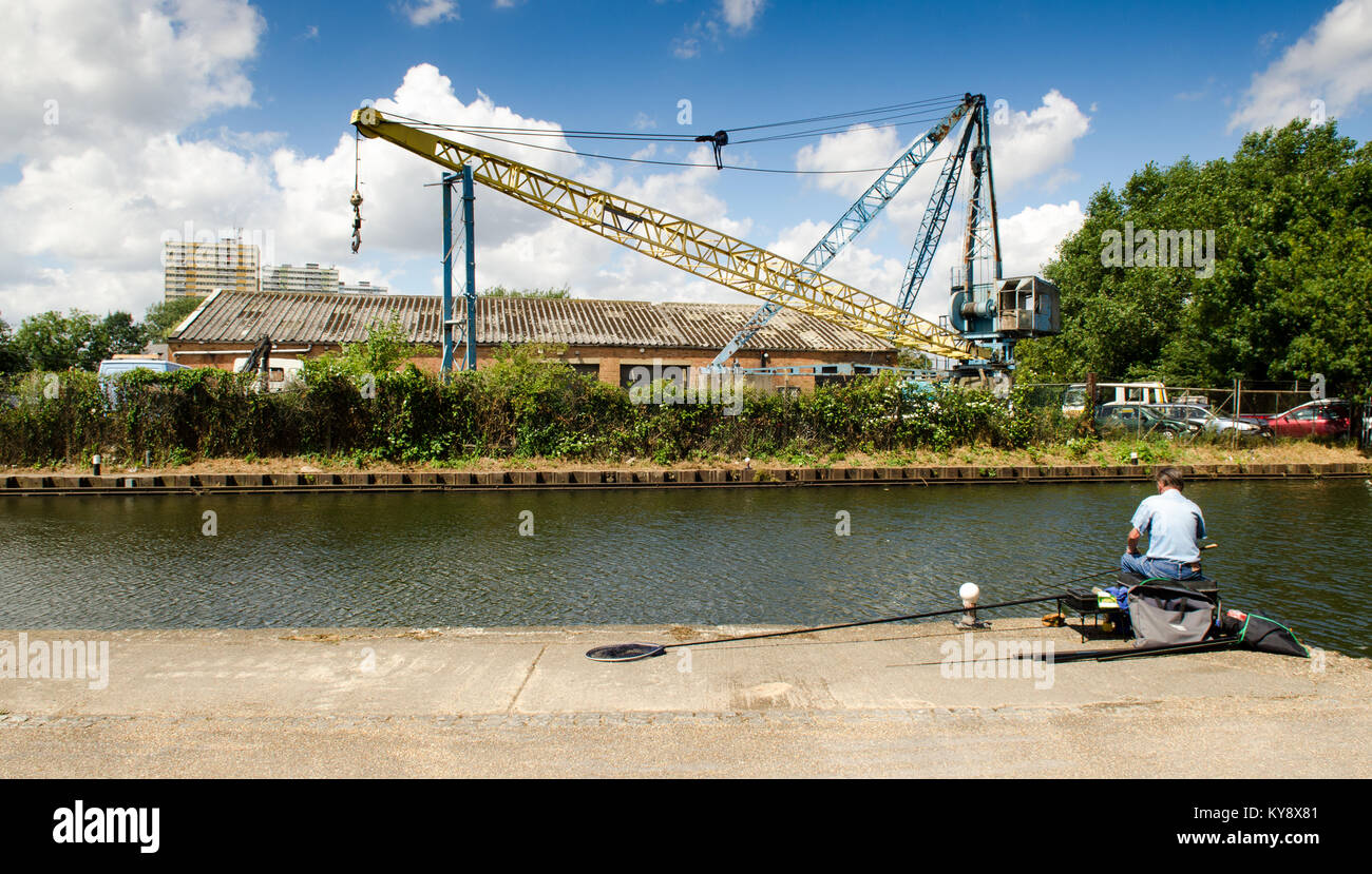 Ein Bewunderer angler sitzt neben dem Kanalisierten Fluss Lea in East London, mit industrial decay und urban high-rise Gehäuse darüber hinaus. Stockfoto