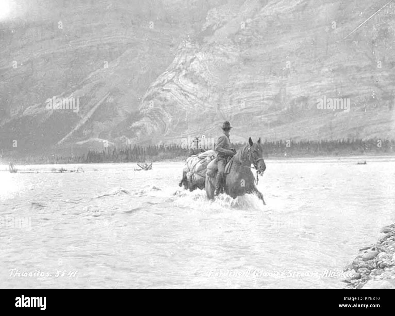Ein Schwarzweißfoto von 1912 von zwei Reitern, die einen Fluss überqueren, in einer ländlichen Landschaft, das Reisen und Arbeiten von Pferden veranschaulicht Stockfoto
