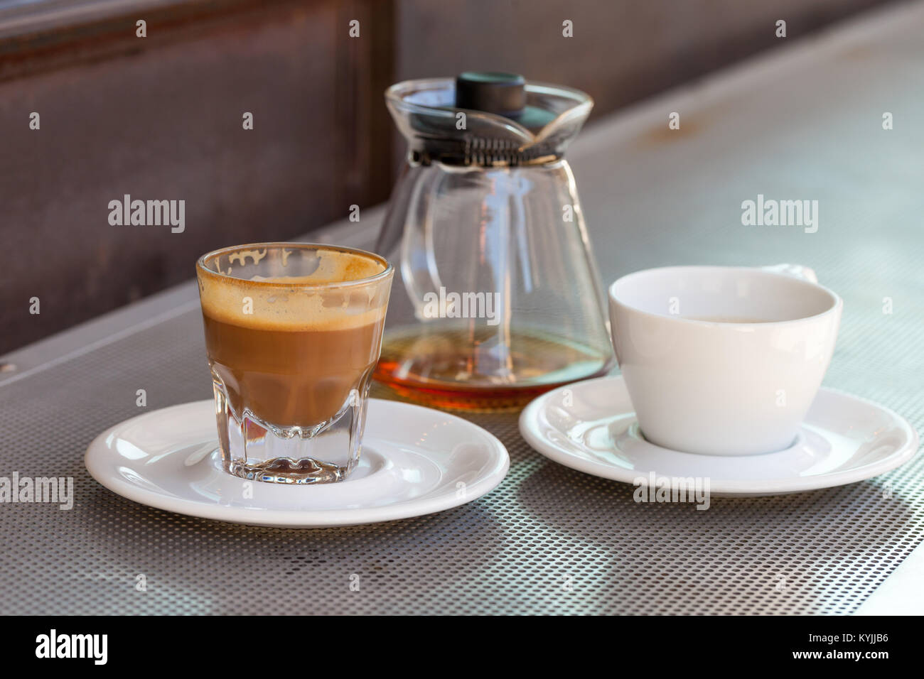 Verbrauchte cortado Kaffee in ein Glas mit Keramik weiße Tasse und Kännchen auf metallischen Tabelle und eine rostige Hintergrund Stockfoto