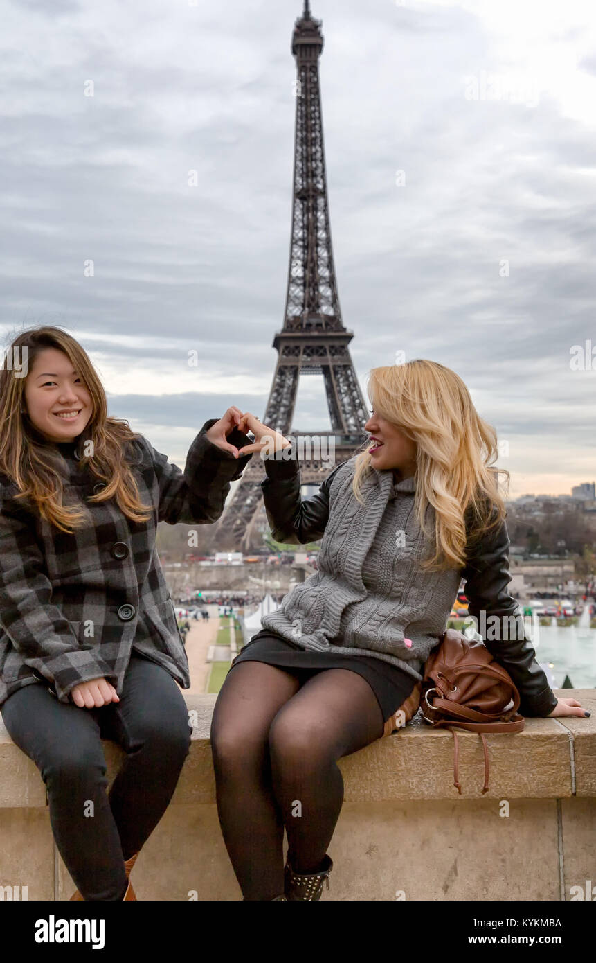Paris Eiffel Tower Touristen posieren für Fotos mit dem Turm im Hintergrund. Zwei lächelnde junge Frauen bilden eine Herzform mit ihren Händen. Stockfoto