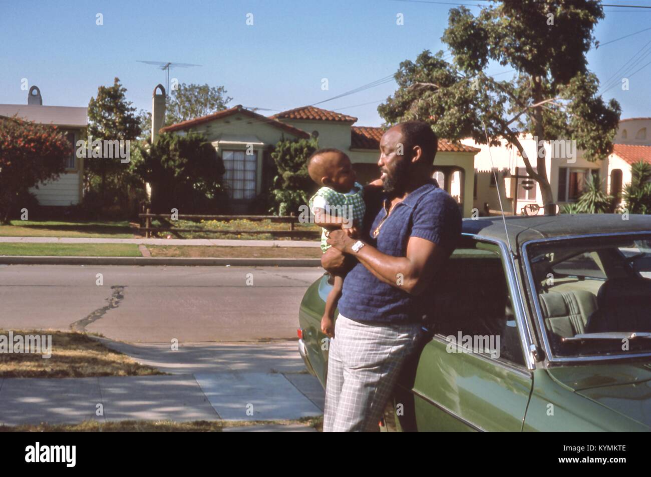 Portrait einer afrikanischen amerikanischen Mann, der an einem afrikanischen amerikanischen Kind er in seinen Armen hält, Juli 1975 lächelnd. Der Mann lehnt sich gegen ein Auto in einer Einfahrt in einer Wohnstraße in Los Angeles, Kalifornien geparkt. () Stockfoto