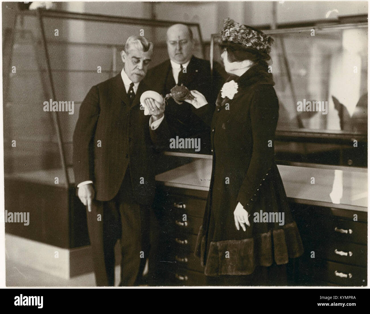 Historische Illustration mit Männern und einer Frau in zeitgemäßer Mode, die vor einem Gehäuse stehen, das Teil des National Museum of the American Indian Collection ist. Stockfoto