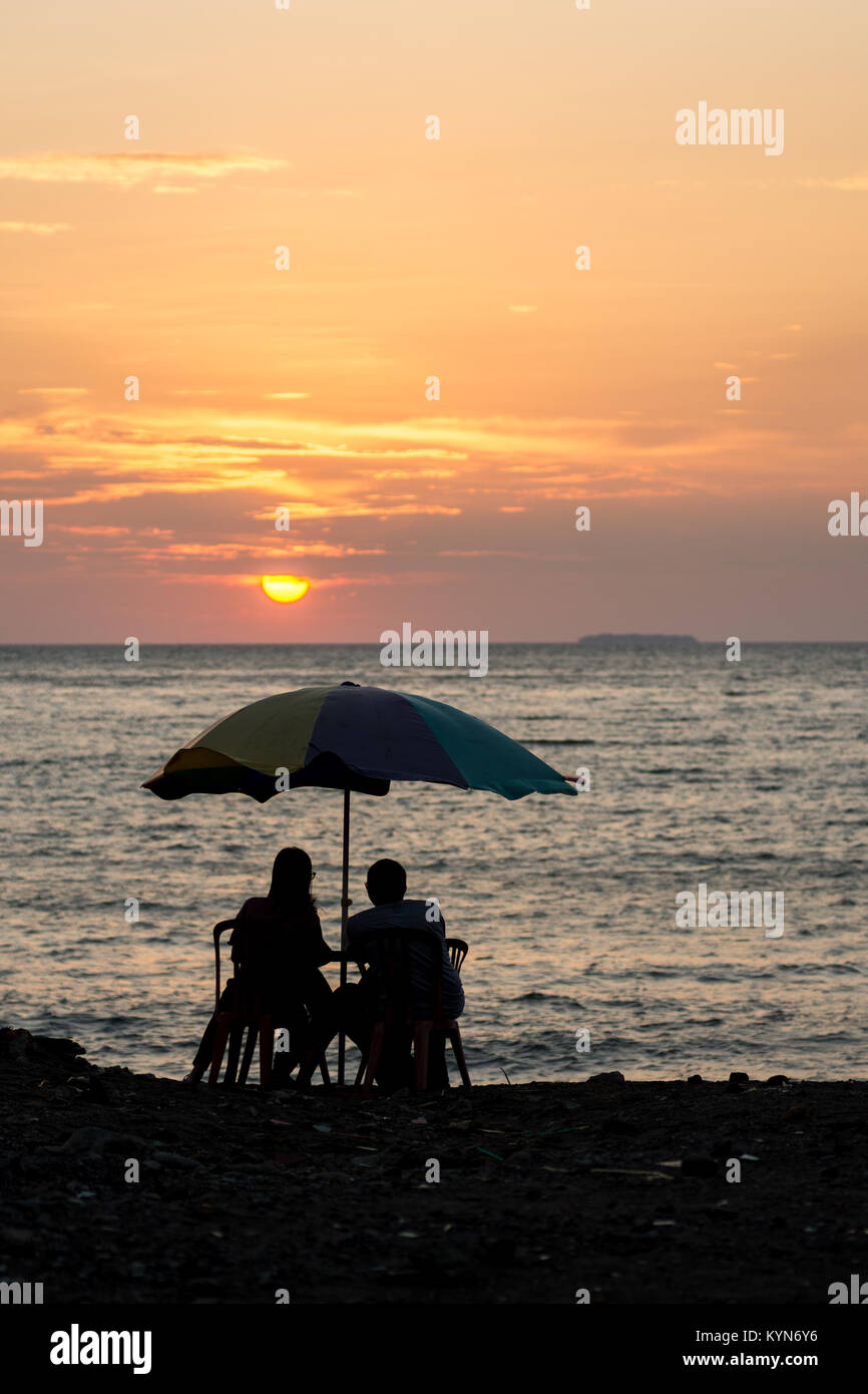 Ein paar romantische Silhouette sitzen an einem grundlegenden Terrassentisch mit Regenbogen Sonnenschirm den Sonnenuntergang auf See mit einer Insel am Horizont. Stockfoto