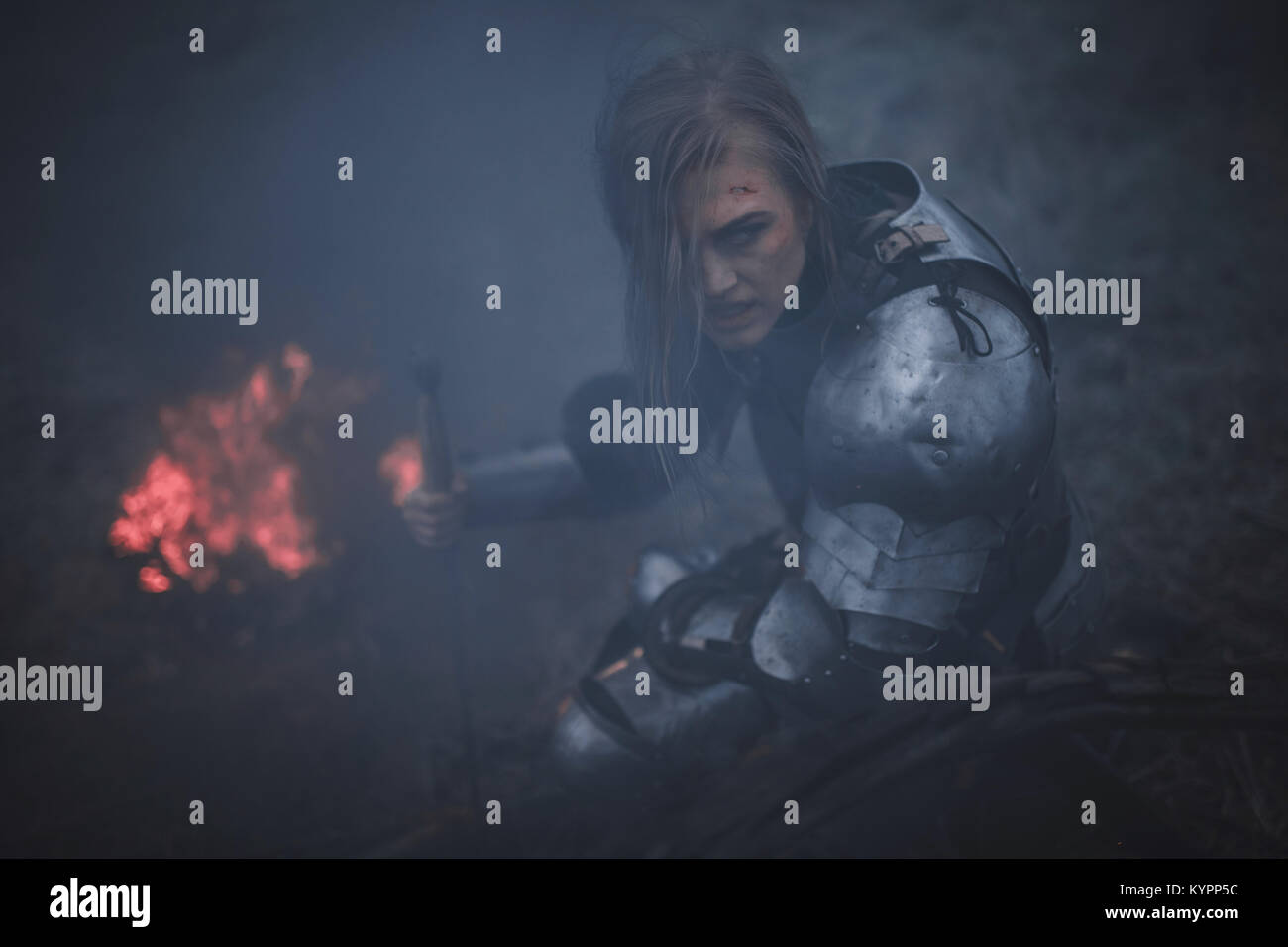 Mädchen in Bild von Jeanne d'Arc in Rüstung und mit dem Schwert in den Händen kniet vor Hintergrund der Feuer- und schwarzen Rauch. Nahaufnahme. Stockfoto
