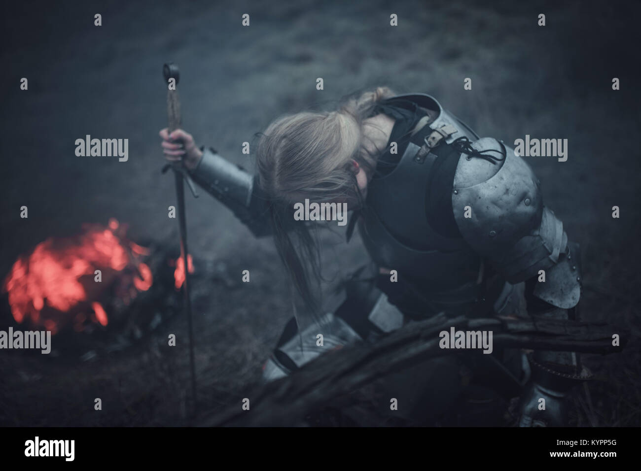 Mädchen in Bild von Jeanne d'Arc in Rüstung und mit dem Schwert in den Händen kniet vor Hintergrund der Feuer- und schwarzen Rauch. Nahaufnahme. Stockfoto