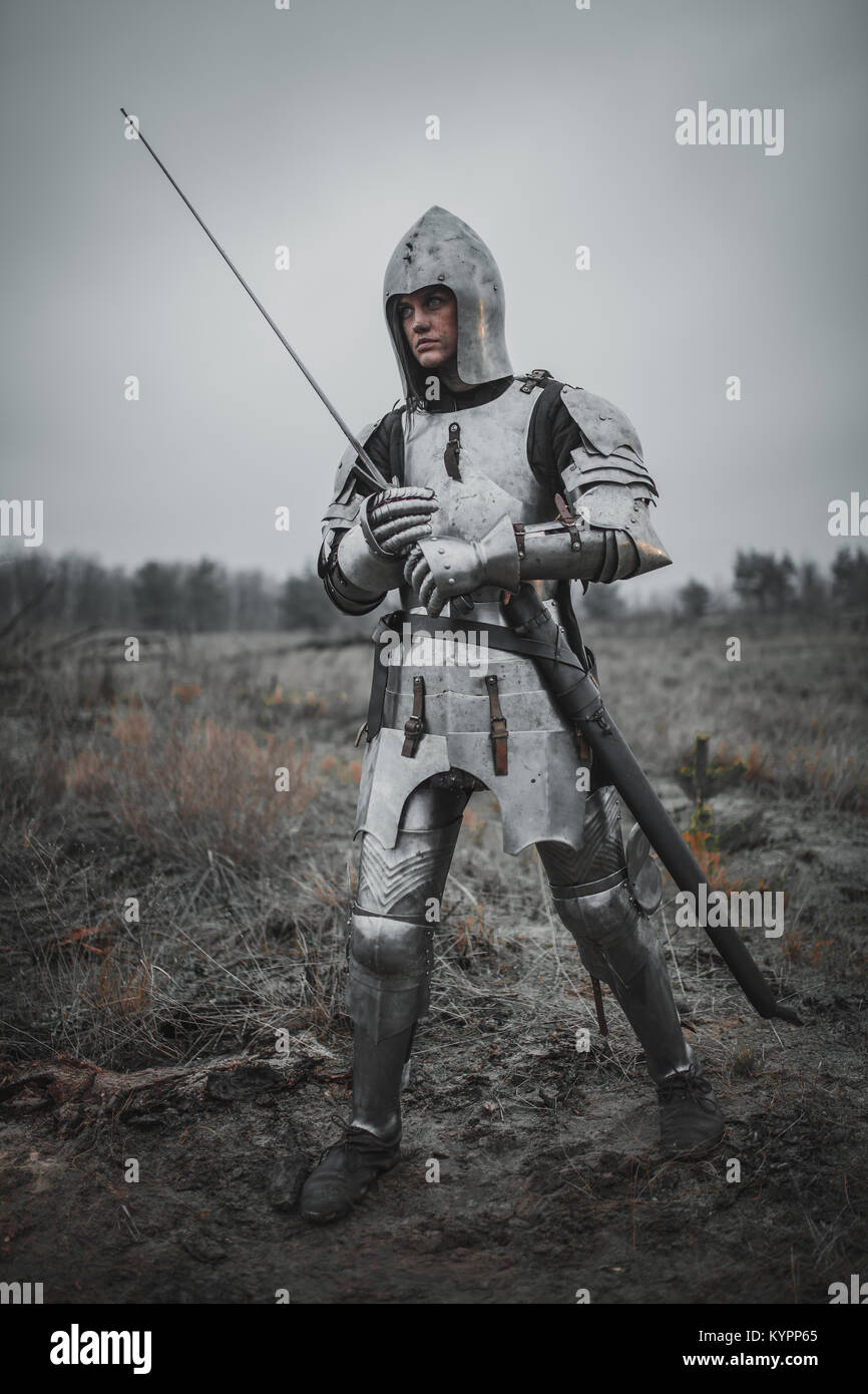 Mädchen in Bild von Jeanne d'Arc geht auf Wiese über Schlamm in Rüstung und Helm mit einem Schwert in den Händen. Stockfoto