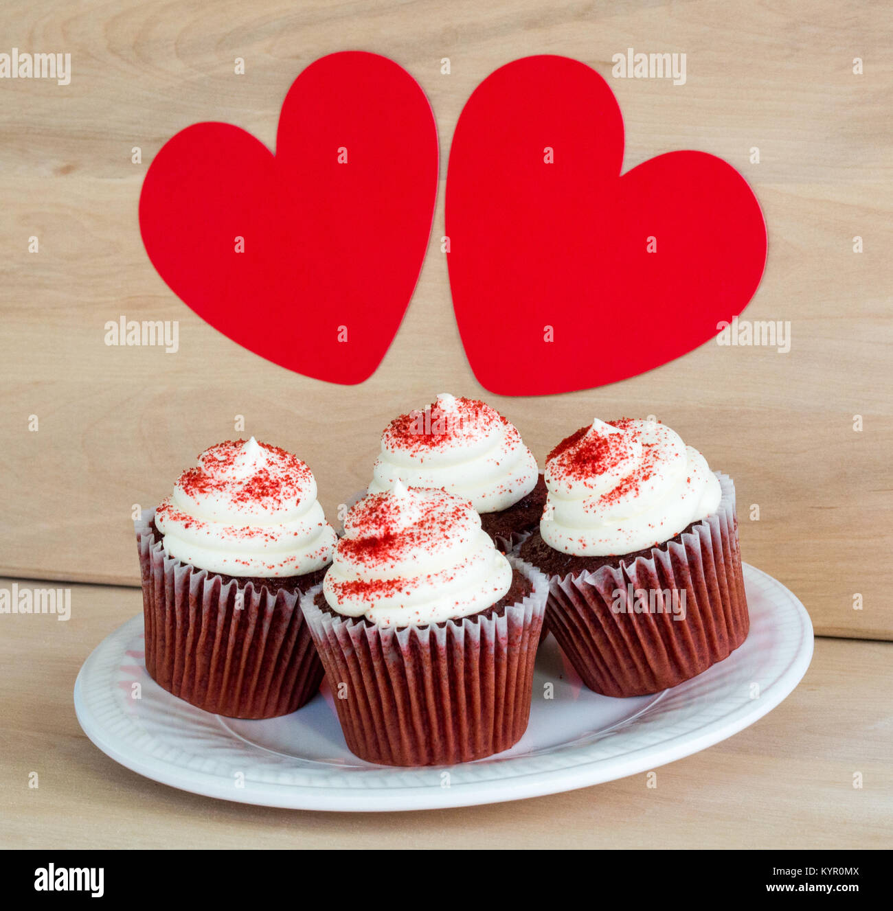 4 chocolate Cupcakes mit weißem Zuckerguss und roten Streusel auf einem Holztisch unter 2 rote Herzen zum Valentinstag Stockfoto