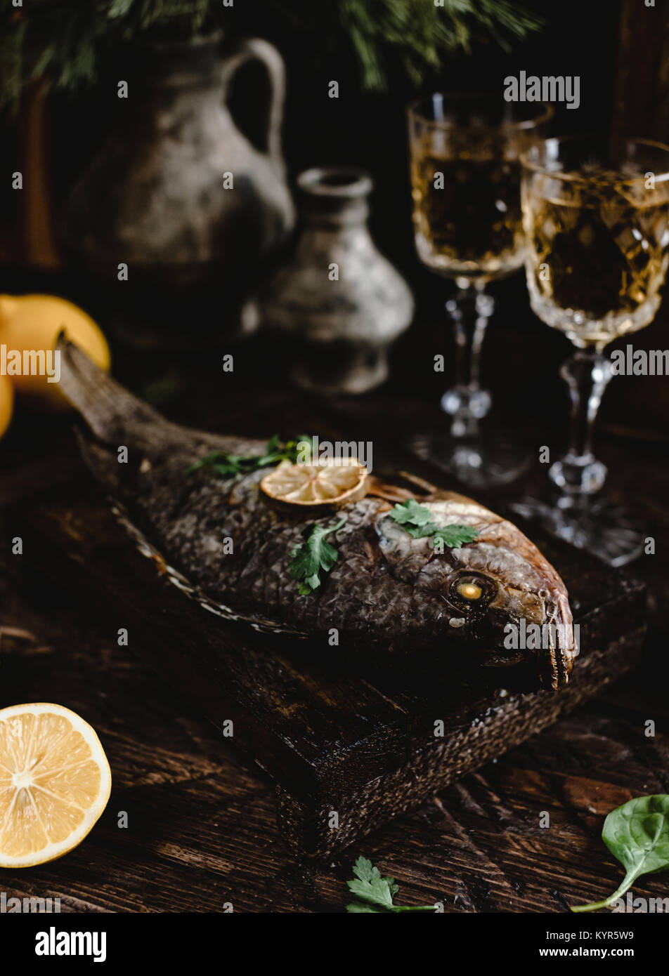 Gebackener Fisch mit Zitrone und Kräutern auf Holzbrett mit weiß Weingläser auf dunklen Holztisch Stockfoto