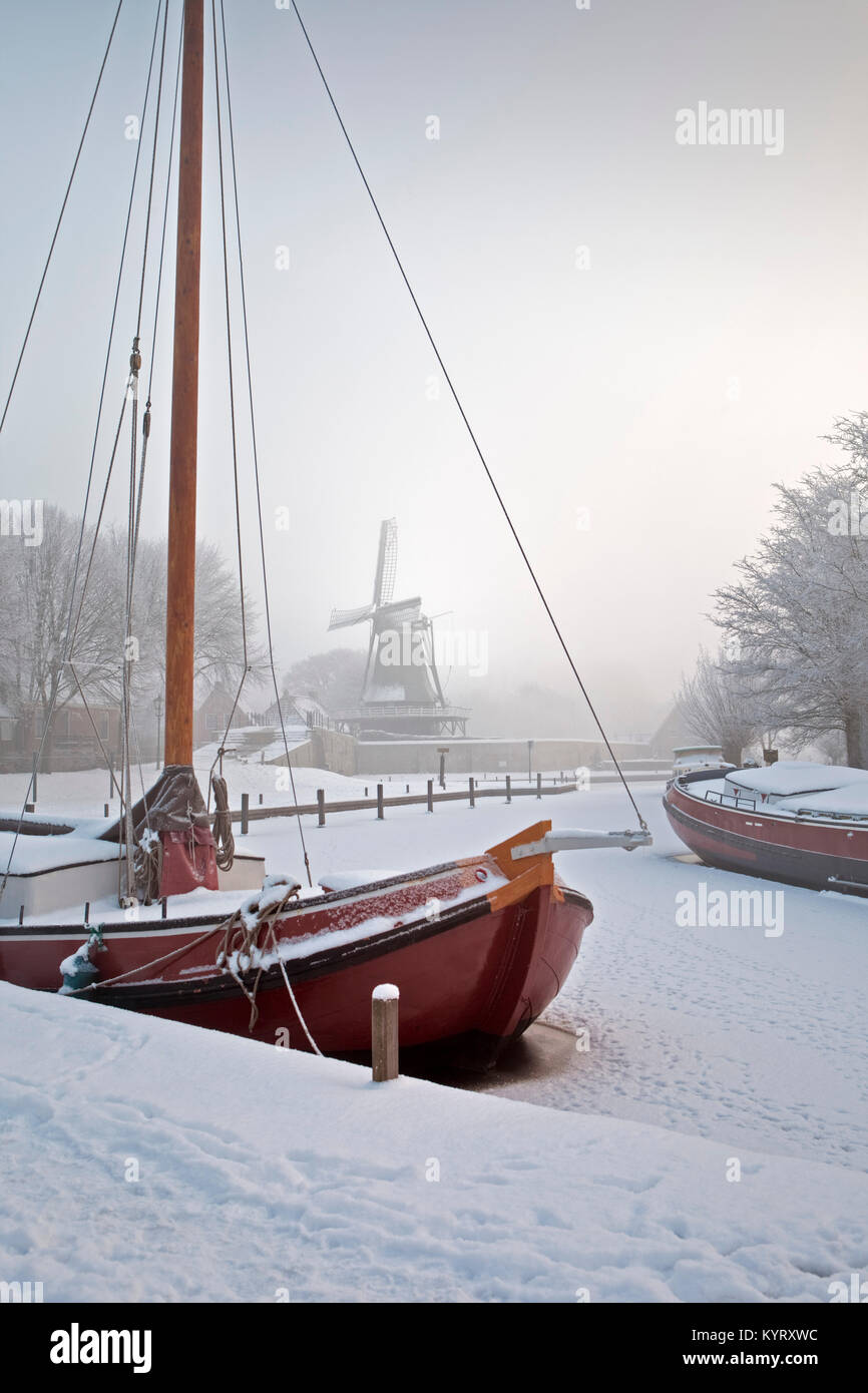 Die Niederlande, Sloten, Traditionelle segeln Frachtschiffe in der gefrorenen Kanal. Hintergrund Windmühle. Stockfoto