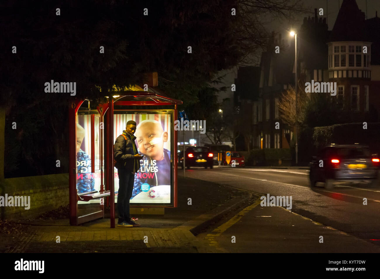 Ein junges Männchen Warten an einer Bushaltestelle auf einem dunklen Winter Abend in Wellingborough Rd, Northampton, Stockfoto