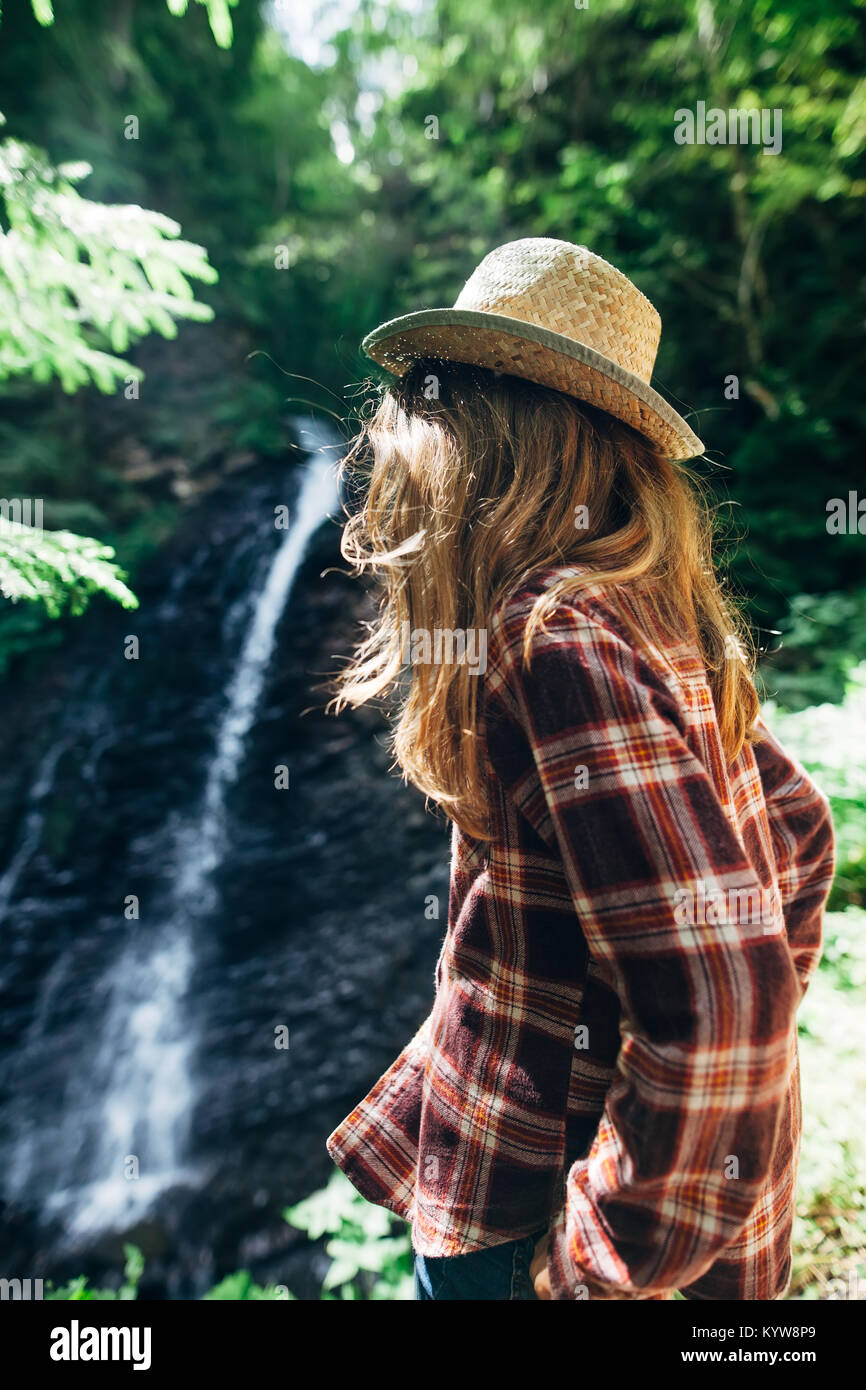 Rückansicht des jungen Mädchens in Shirt und Strohhut aus der Ferne bewundert Wasserfall. Kaukasischen jungen Dame sieht bei fallendem Wasser, Foto auf frische Luft. Porträt Stockfoto