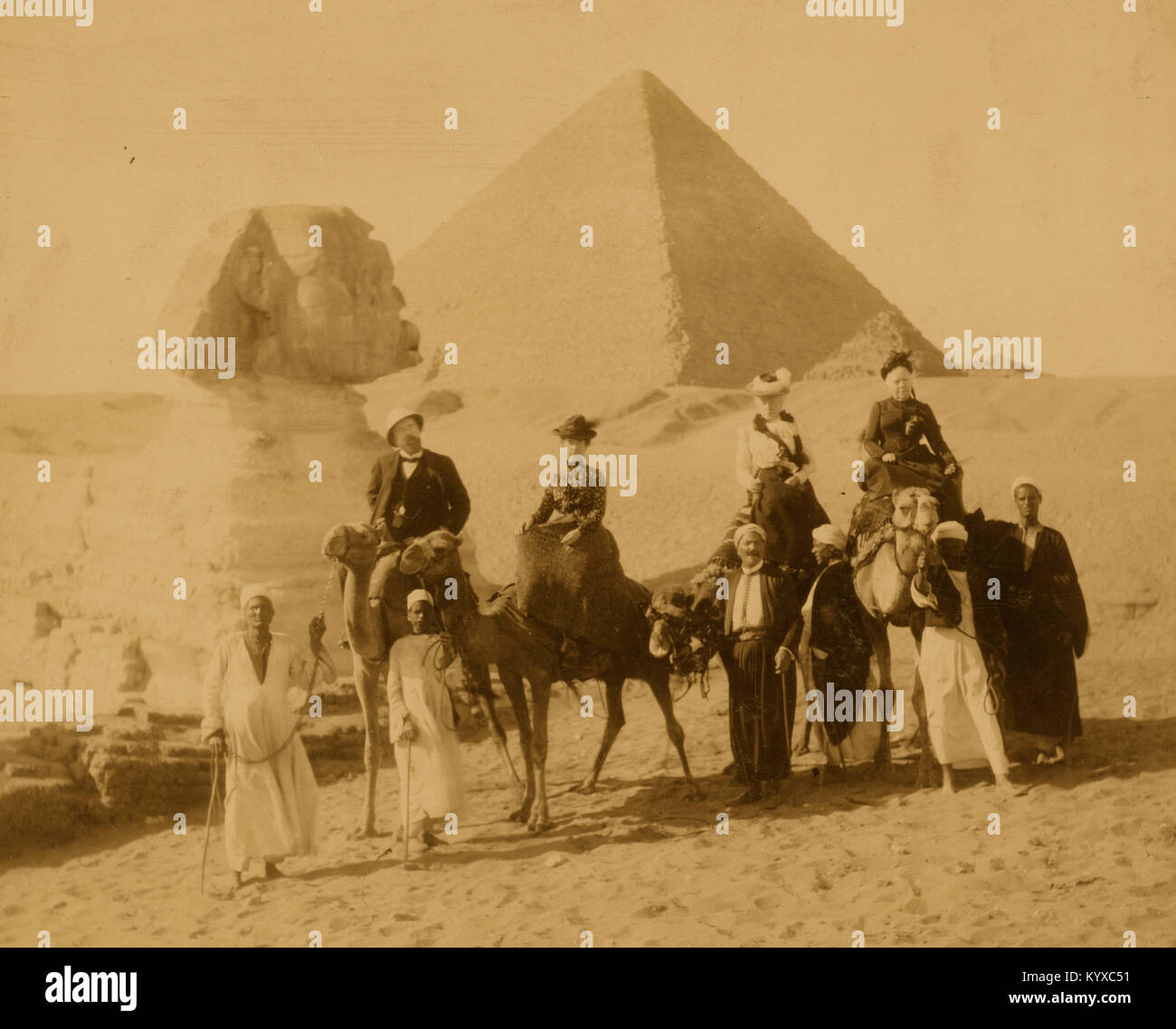 Drei Frauen und ein Mann in westlichen Kleidung, auf Kamelen sitzt, mehrere lokale Männer vor den Kamelen Holding die Zügel, die Pyramiden und die Sphinx im Hintergrund stehen Stockfoto