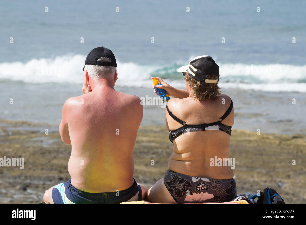 Brirs im Ausland: Frau Anwendung spray Sonnencreme zurück der Mann am Strand in Spanien einen Sonnenbrand zu. Stockfoto