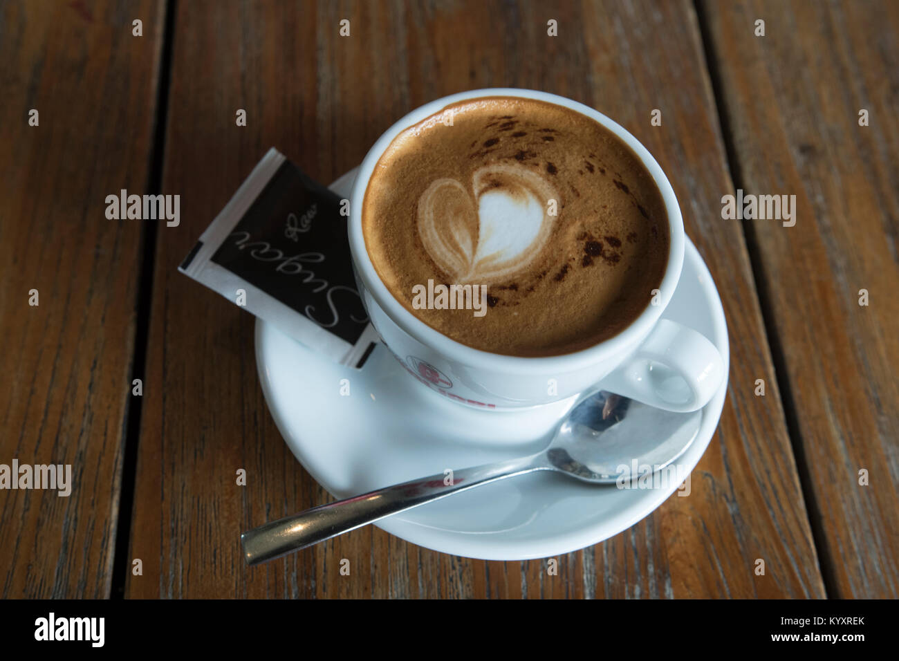 Eine Tasse Cappuccino sitzt auf einem Holzbrett Tabelle. Die Schale ruht in einem weißen Untertasse, mit einem Teelöffel und ein Päckchen Zucker flankiert Stockfoto