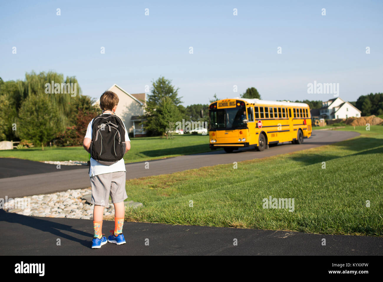 Ansicht der Rückseite des Jungen mit Rucksack warten auf School Bus gegen den klaren Himmel auf Rasen Stockfoto