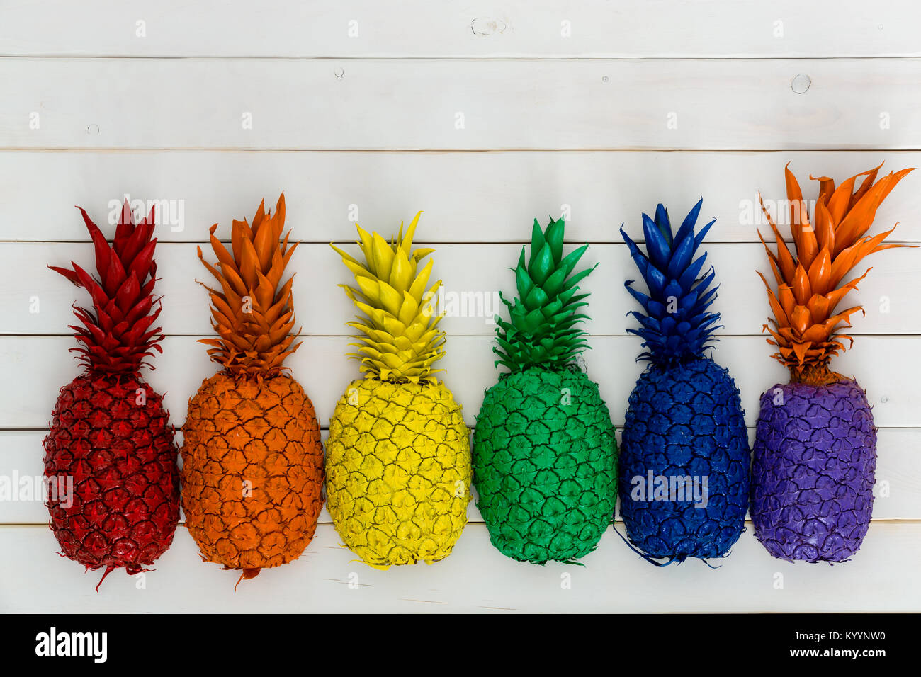 Reihe von bunt leuchtenden Regenbogenfarben Ananas mit ihre Blätter bilden einen unteren Rand auf weißem Holz mit Kopie Raum Stockfoto