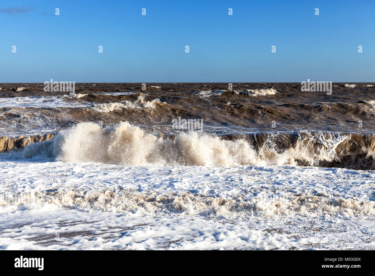 Wellen an der Küste von einem Kiesstrand mit Schaum in den Vordergrund vor einem horizontalen Horizont mit blauem Himmel Stockfoto
