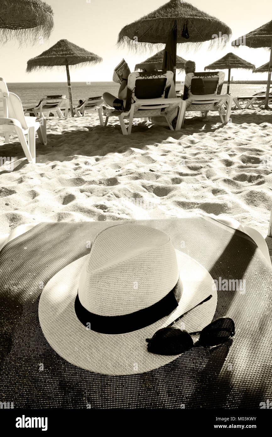 White Panama Sonnenhut und schwarze Sonnenbrille auf ein Handtuch auf die Sonnenliege am Strand an einem sonnigen Tag, im Hintergrund ist ein Sandstrand und Mehr s Stockfoto