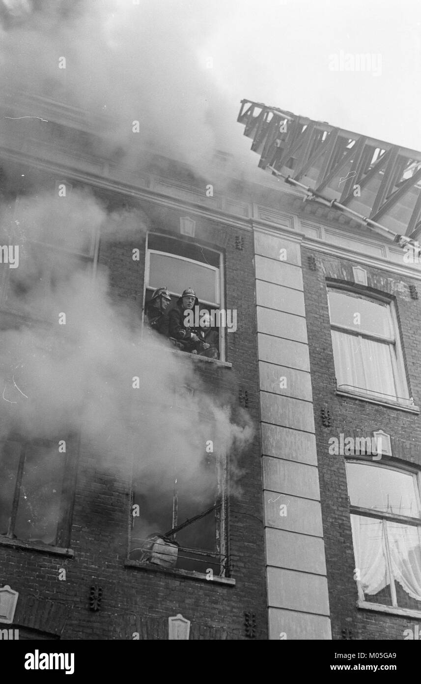 Ein historisches Foto, das ein Feuer in der Straße Jacob van Campenstraat zeigt, auf dem Feuerwehrleute neben einer älteren Frau warten. Das Bild erfasst die Dringlichkeit und Intensität von brandbedingten Notfällen in städtischen Umgebungen. Stockfoto