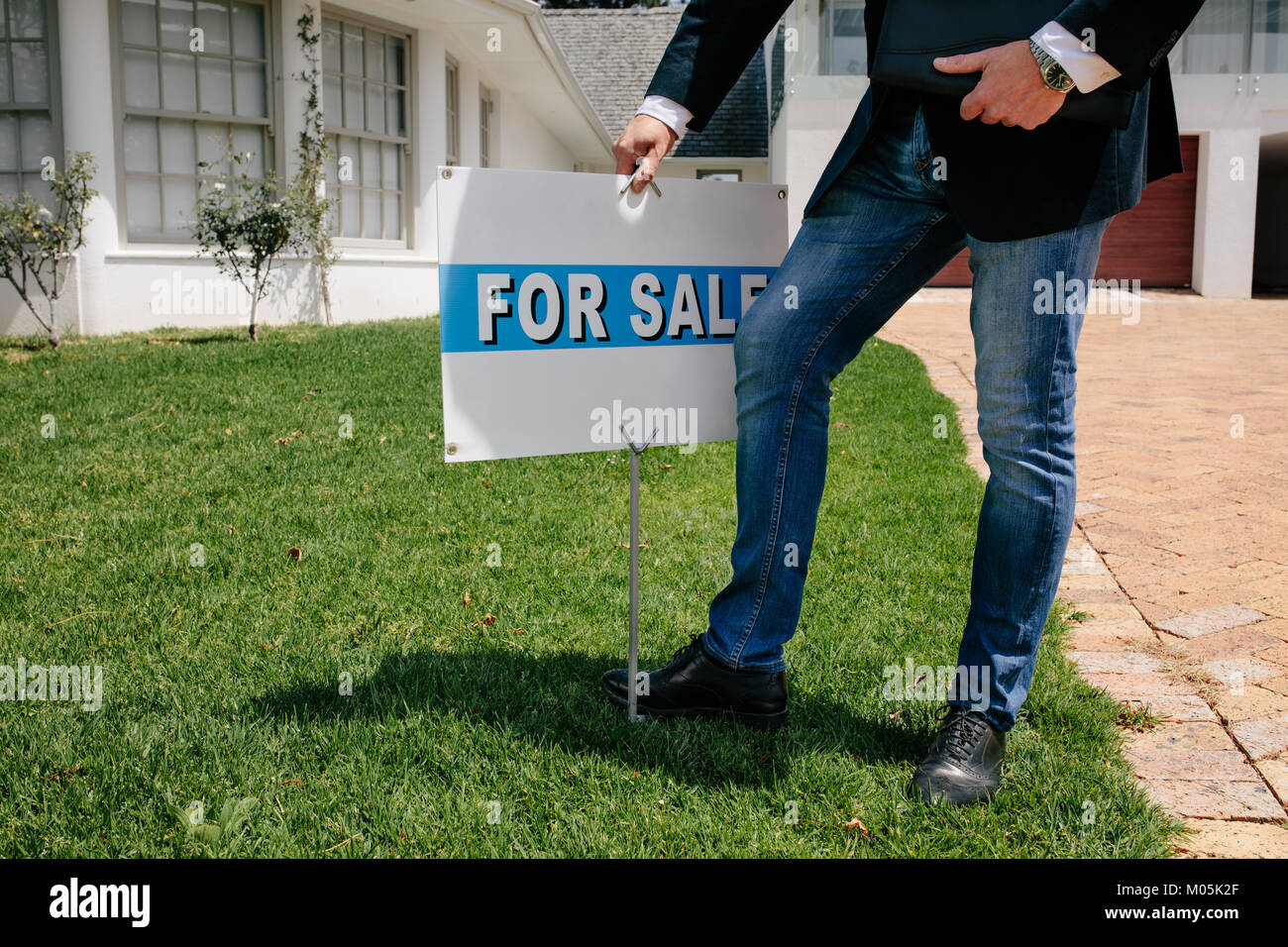 Immobilienmakler mit einem Zu Verkaufen Schild außerhalb eines Hauses. Männliche Real Estate Broker mit zum Verkauf im Rasen von einem neuen Haus. Stockfoto