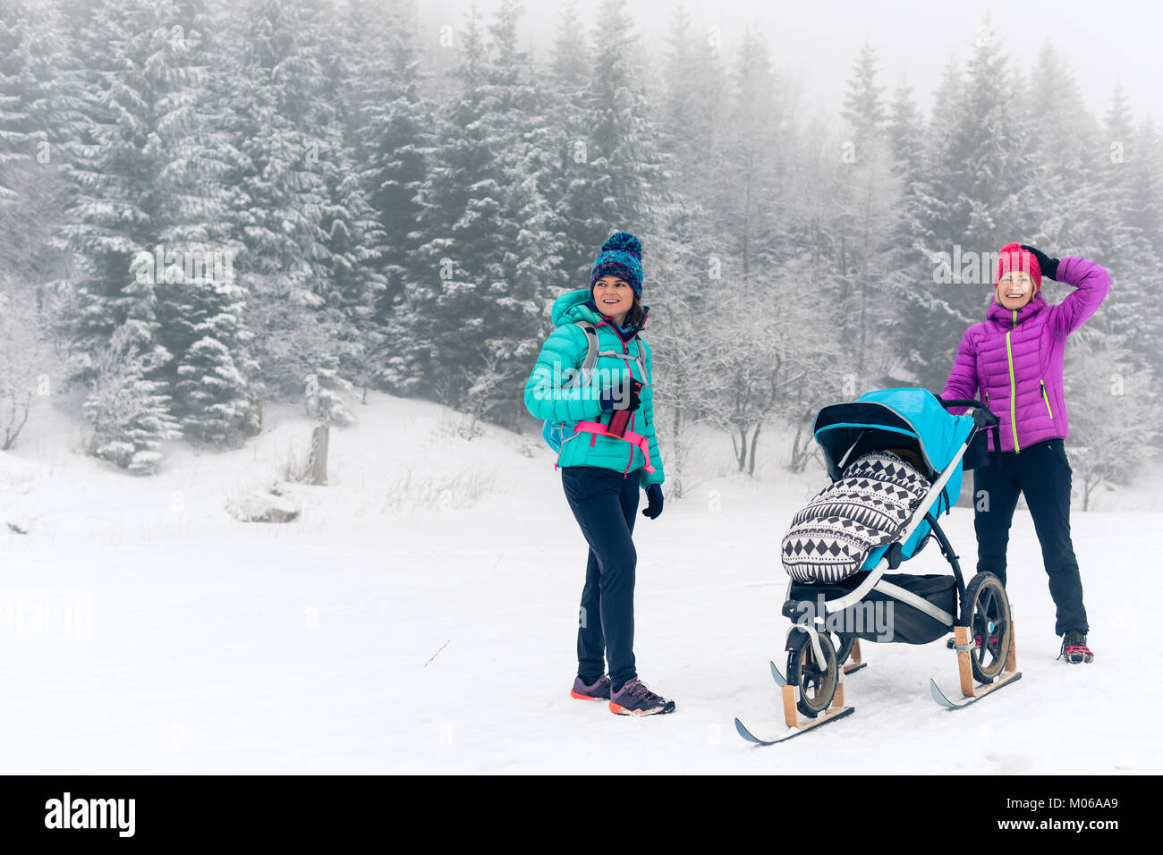 Mutter mit Baby Stroller genießen Mutterschaft im Winter Wald mit Freundin oder Partner. Wandern oder Walken Frau mit Schlitten pram in Wäldern. Stockfoto